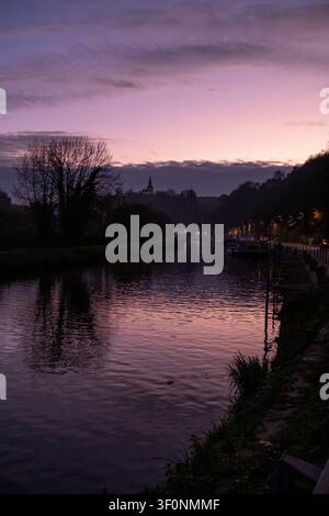 Vista del fiume Rance al crepuscolo con silouette di alberi scuri, moli, cielo al tramonto viola-rosa e un campanile lontano vicino al porto di Dinan a Dinan Foto Stock