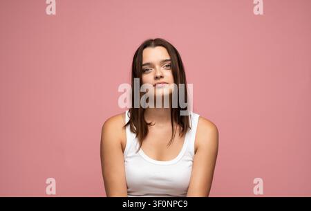 Giovane donna che posa con sicurezza in una semplice canottiera bianca con capelli naturali e un'espressione facciale seria su sfondo rosa Foto Stock
