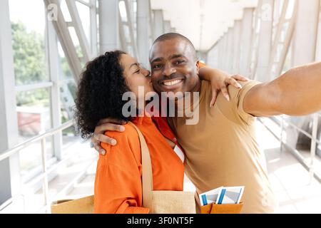 Donna nera che bacia la guancia del fidanzato mentre scatta un selfie in aeroporto Foto Stock