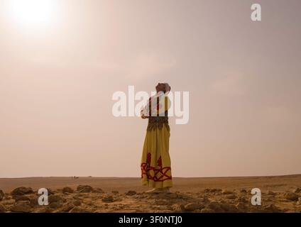 Donna solitaria che indossa un costume tradizionale ricamato giallo rosso vivace, con le braccia incrociate in autoabbraccio, che guarda verso l'alto nel cielo deserto luminoso e nebbioso Foto Stock
