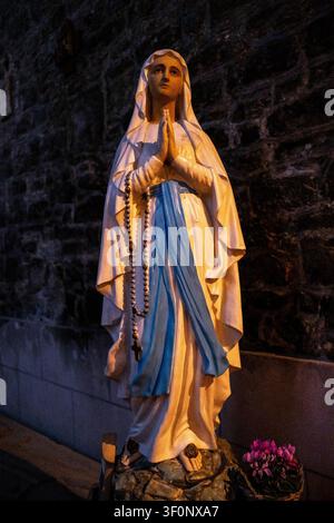 Vista di una statua della Vergine Maria, nota come Nostra Signora di Lourdes, con abiti bianchi, fascia blu, mani in preghiera, rosario e fiori su uno sfondo di muro di pietra Foto Stock