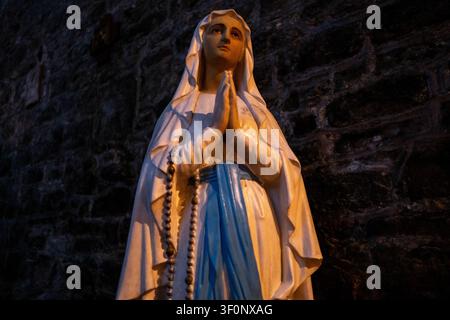 Vista di una statua della Vergine Maria, nota come Nostra Signora di Lourdes, con abiti bianchi, fascia blu, mani in preghiera, rosario e fiori su uno sfondo di muro di pietra Foto Stock
