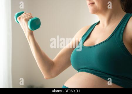 Una donna incinta è in piedi tenendo un manubrio in una posa rilassata e casuale contro un muro bianco. La madre e atleta mostra movimenti dolci e naturali di cura personale con una sicurezza calma e bilanciata al chiuso. Foto Stock
