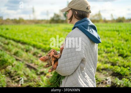 Una donna e un agricoltore tengono carote appena raccolte, guardando lontano attraverso un campo da dietro. La scena calma e contemplativa rivela prodotti biologici naturali e radicati nella campagna. Foto Stock