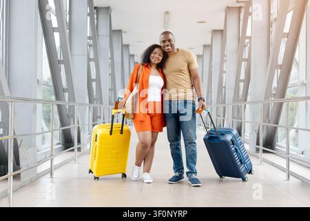 Giovane coppia nera sorridente in piedi insieme in un corridoio luminoso dell'aeroporto con le valigie Foto Stock