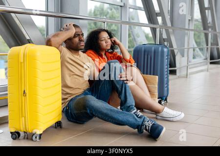 Giovane coppia nera seduta sul pavimento del corridoio dell'aeroporto accanto alle valigie, con aria stanca Foto Stock
