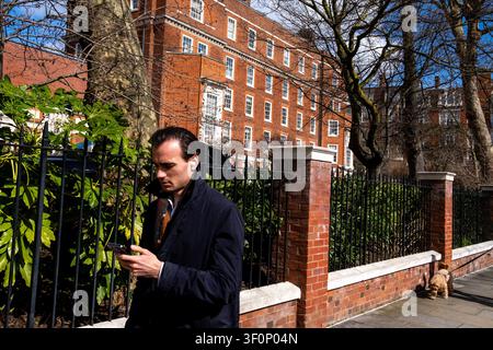 Un uomo guarda uno smartphone con le cuffie su un marciapiede tranquillo a Kensington, con un guardiano di cani e edifici residenziali di lusso in mattoni rossi dietro inferriate di ferro Foto Stock