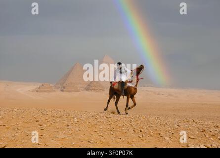 Cavaliere arabo trionfante in camicia bianca e keffiyeh che galoppa attraverso la sabbia dorata del deserto davanti alle iconiche piramidi di Giza sotto un vivido arco di arcobaleno Foto Stock