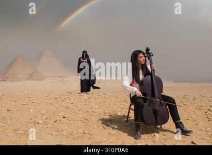 Violoncellista donna seduta che suona il violoncello alle iconiche piramidi di Giza con una donna vestita in modo tradizionale e un vivido arco di arcobaleno in un drammatico cielo desertico, ideale per Foto Stock