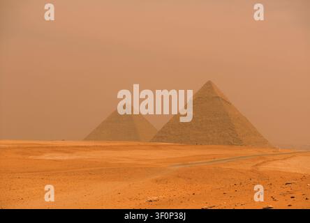 Piramidi di Giza avvolte in una drammatica foschia di tempesta di sabbia arancione sopra il vasto deserto del Sahara, ripresa ampia ideale per editoriali di viaggio in Egitto e narrazioni sul clima Foto Stock