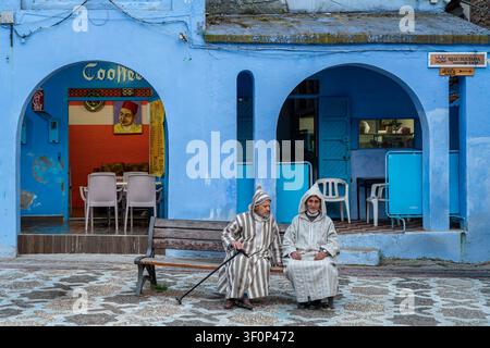 due uomini locali vestiti con una burnus tradizionale sono seduti su una panchina davanti a un caffè dipinto di blu con porte ad arco, incarnando la vita quotidiana di Chefc Foto Stock
