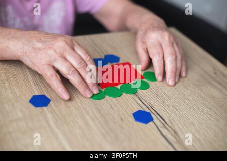Prima piano ritagliato di un ritratto di una donna anziana che raccoglie figure geometriche di legno a casa seduta al tavolo. Allenamento della memoria per la prevenzione della demenza Foto Stock