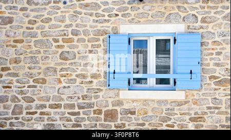 Primo piano di un muro di pietra rustico con una finestra con cornice bianca dotata di persiane in legno azzurro e una barra trasversale, che mostra una tradizionale a mediterranea Foto Stock