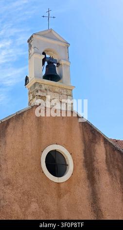 Primo piano di un campanile rustico con una campana in metallo e una croce in cima, caratterizzato da una finestra rotonda e da un esterno ruvido sotto un cielo blu terso. Foto Stock