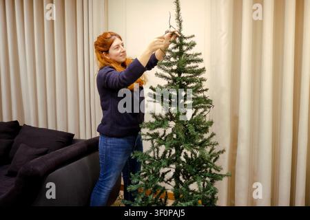 Donna che appende luci natalizie a un albero artificiale verde, celebrando le feste invernali a casa Foto Stock