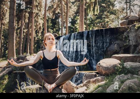 Giovane donna seduta in posizione yoga all'aperto vicino a una cascata. Donna pratica yoga all'aperto in una radura del bosco. Ritiro di detox digitale, salute mentale Foto Stock