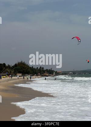 Kitesurfer che cavalcano onde oceaniche ventose vicino a una spiaggia sabbiosa tropicale. Persone attive si divertono con il kitesurf su un mare ondulato sotto un cielo nebbioso. Turisti passeggiano lungo Foto Stock