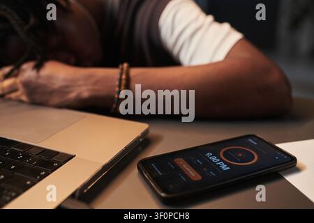 Uomo nero che riposa accanto a un laptop e all'allarme del telefono durante un esaurimento lavorativo Foto Stock