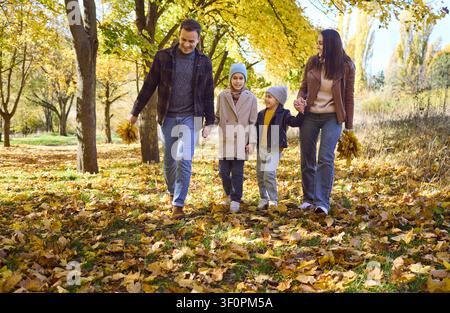 Famiglia felice con due bambini che cammina nel parco autunnale, tenendosi per mano e sorridendo Foto Stock