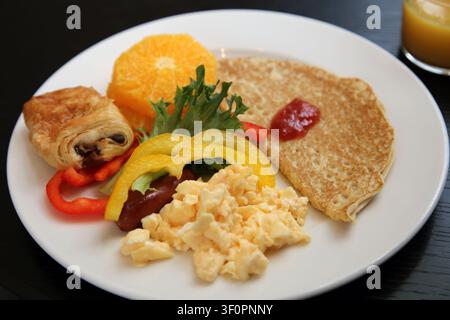 Deliziosa colazione nordica in hotel che include uova strapazzate, verdure, crepe con marmellata di frutti di bosco, frutta fresca, croissant al cioccolato e succo d'arancia. Foto Stock
