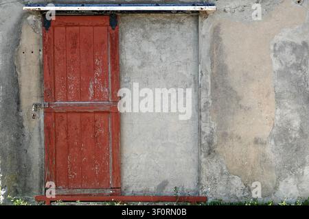 Porta di legno rossa con assi verticali, leggermente consumata, con vernice scrostata. È dotata di cerniere metalliche ed è inserita in un muro di cemento ruvido. Foto Stock
