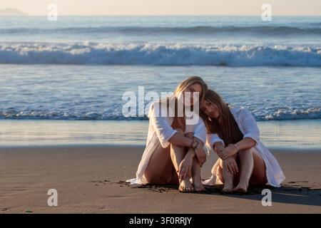 Due giovani donne sorridenti sedute sulla sabbia, godendosi le onde dell'oceano all'ora dorata Foto Stock