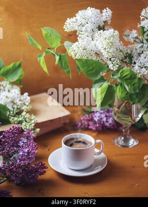 Sfondo romantico per la colazione. Una tazza di caffè espresso fragrante su un tavolo di legno con un mazzo di lillà bianchi e viola e un libro Foto Stock