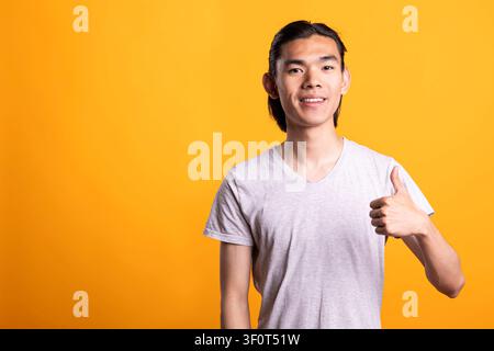 Uomo affascinante si trova con sicurezza su uno sfondo giallo, sorridendo e facendo il gesto del pollice alzato. Giovane modello maschile in abbigliamento casual mostra approvazione, felicità e successo con un'espressione facciale positiva Foto Stock