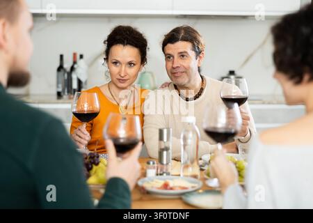 Un gruppo di persone gioiose festeggia una vacanza, sorseggiando un drink e chiacchierando intorno al tavolo della propria casa Foto Stock