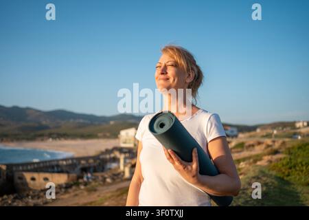 Felice donna caucasica anziana in occhiali con tappetino da yoga sorridente sulla riva dell'oceano. Donna in pensione che gode di viste sulla natura prima dello yoga mattutino. Sentire armonia. Concetto di stile di vita sano e anziano Foto Stock