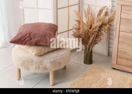 Interno accogliente della casa con cuscini di velluto a coste impilati su un pouf soffice, erba di pampas essiccata in un vaso e un cassettone in legno Foto Stock