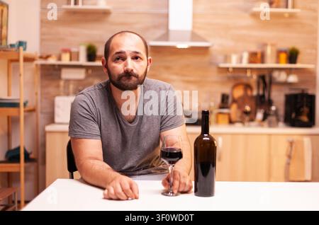 Giovane uomo pensieroso in un ambiente domestico che tiene un bicchiere di vino rosso e riflette sui problemi della vita. Persona infelice con sensazioni di malattia e ansia, esausta per problemi di alcolismo. Foto Stock