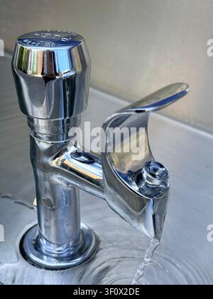 Primo piano dell'acqua del rubinetto che scorre da un rubinetto cromato in un lavandino in acciaio inossidabile, illustrando l'uso dell'acqua potabile, la conservazione e la scarsità idrica. Foto Stock