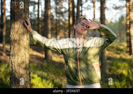 Escursionista gioiosa che si gode una passeggiata nel bosco e prova emozioni positive, in piedi accanto a un albero sorridendo. Foto Stock