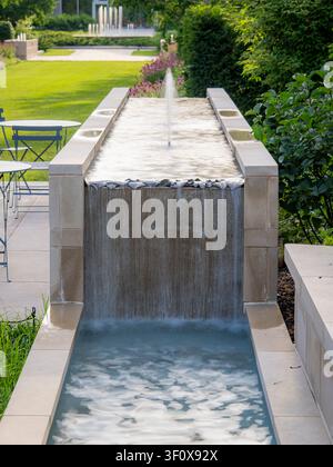 Fontana moderna in pietra con un getto centrale e una cascata a cascata che sfocia in una piscina da giardino pubblico in una giornata estiva soleggiata Foto Stock
