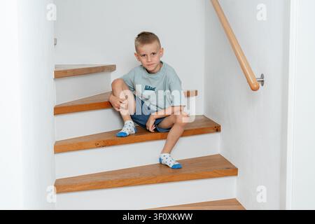 Ragazzo seduto sulle scale, in scala interna con pareti bianche e ringhiera in legno, con gradini, ringhiera in legno, parete bianca, con legno, texture di intonaco Foto Stock