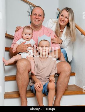 Un ritratto di famiglia di un uomo, una donna, una bambina e un bambino con una corona seduti insieme su una scala di legno con un corrimano in tessuto morbido, catturato in una luce calda Foto Stock