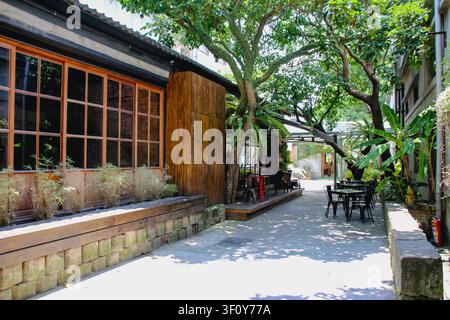 Sentiero esterno panoramico di un edificio storico con finestre in legno e tavoli da caffè sotto alberi verdi. Foto Stock