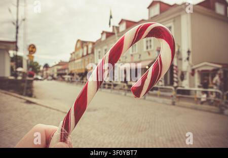 Prima vista ravvicinata di una mano di turista che tiene Polkagris rossi e bianchi, caramelle svedesi a bastoncino, con la città di Gränna sfocata sullo sfondo in Svezia. Foto Stock