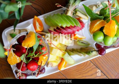 Un vassoio bianco contiene vari frutti come fragole, arance, mele e uva. I frutti sono disposti in bicchieri e sparsi intorno al vassoio. Foto Stock