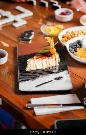 Una fetta di dessert è su un piatto nero con una guarnizione decorativa. Intorno al piatto ci sono varie ciotole con frutta, snack e bevande. Le persone si godono Foto Stock