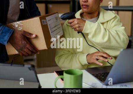 Donna biracial di mezza età che scansiona l'etichetta di spedizione su un pacco mentre un collega tiene una scatola vicino al laptop, elaborando l'adempimento dell'ordine. Ideale per e-commerce, flusso di magazzino, servizio di consegna Foto Stock