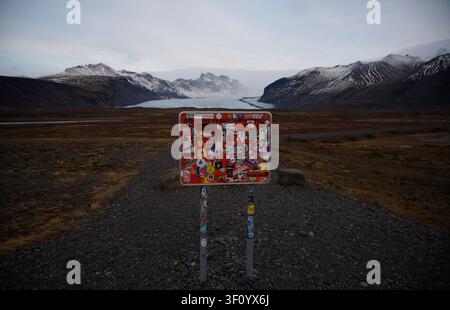 Un cartello di chiusura della strada ricoperto di adesivi si erge su un sentiero di ghiaia scura davanti alla lingua sporgente del ghiacciaio Skaftafellsjökull Foto Stock