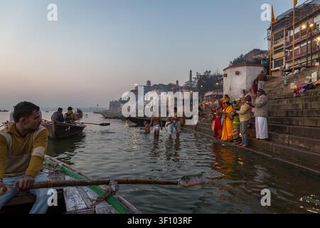 Barcaiolo che rema oltre i pellegrini che eseguono rituali ai ghati di Varanasi sul fiume Gange al crepuscolo, Uttar Pradesh, India. Foto Stock