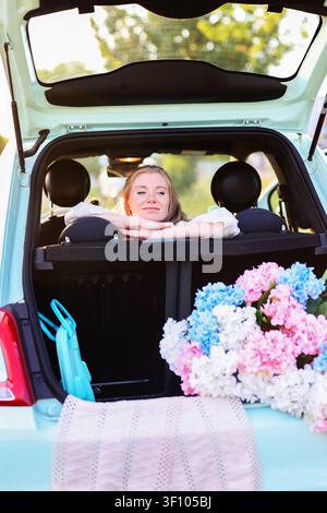 Concetto di vacanza estiva con una donna sorridente, bagagliaio aperto, fiori colorati e luce calda in un paesaggio di campagna scenografico Foto Stock