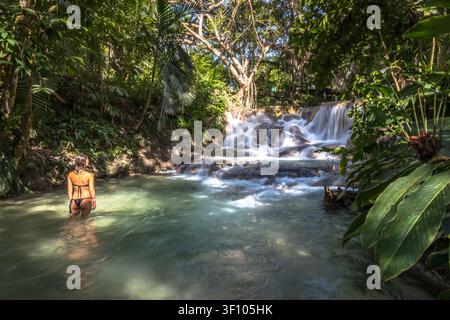 Una turista in bikini che guada i bacini naturali delle Dunn's River Falls, circondata dalla lussureggiante giungla tropicale a Ocho Rios, Giamaica. Foto Stock