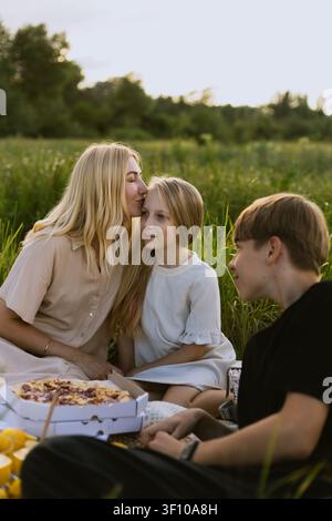 Una madre amorevole condivide un momento tenero con sua figlia mentre gode di un picnic con suo figlio in un prato illuminato dal sole Foto Stock