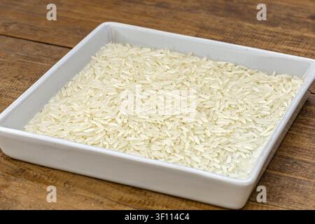 Foto ravvicinata di cibo di riso al gelsomino thailandese in una ciotola quadrata bianca su legno rustico, evidenziando i chicchi di riso al gelsomino bianco e marrone Foto Stock