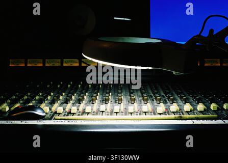 Vista laterale di una lente d'ingrandimento illuminata che illumina i fader su una console di mixaggio audio vintage, catturando un'atmosfera autentica da studio analogico degli anni '80. Foto Stock