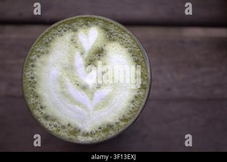Vista dall'alto del tè verde matcha con una bellissima arte matcha servita in un bicchiere e su un sottobicchiere di legno con lo sfondo sfocato di un tavolo di legno Foto Stock